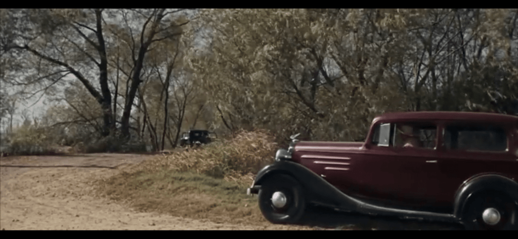 A classic vintage car parked on a dirt road, surrounded by trees and grass, suggesting a scene from a film or countryside setting.