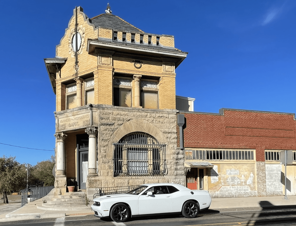 A historic bank building with 'Farmers and Merchants Bank' inscribed above the entrance. A white car is parked in front of the building under a clear blue sky.