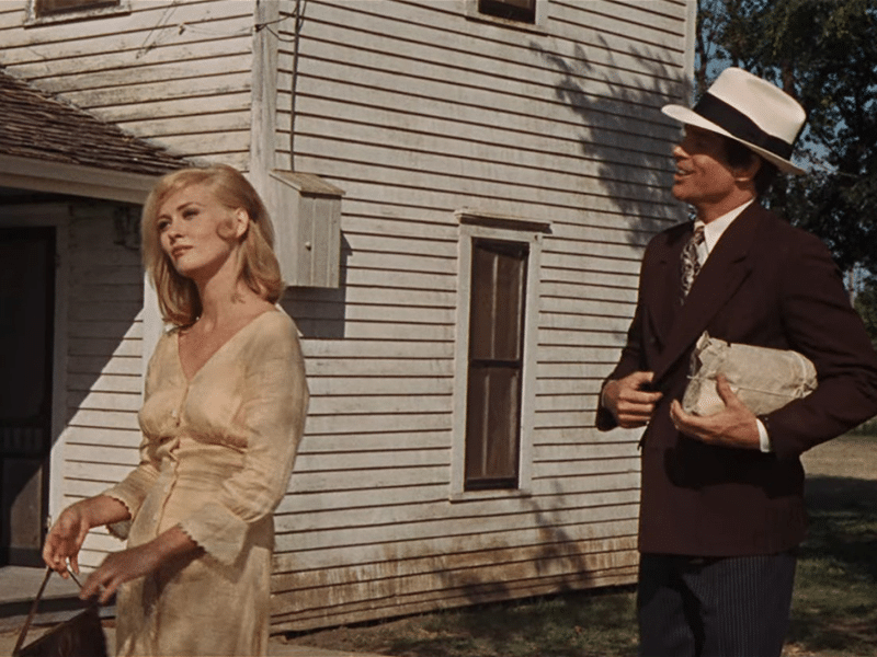 Scene from the film 'Bonnie and Clyde' featuring a woman in a light dress and a man in a suit and hat standing outside a white wooden house.