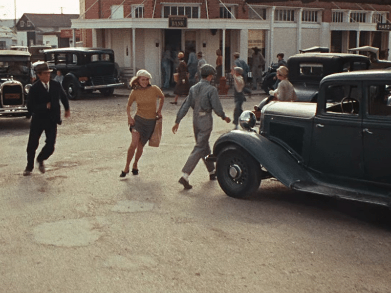 A scene from 'Bonnie and Clyde' showing a bank robbery in a small town, with people running away and vintage cars parked nearby.