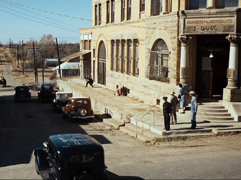 Exterior view of the Farmers and Merchants Bank, featuring a vintage car parked in front and several people in period-appropriate attire on the steps and sidewalk.