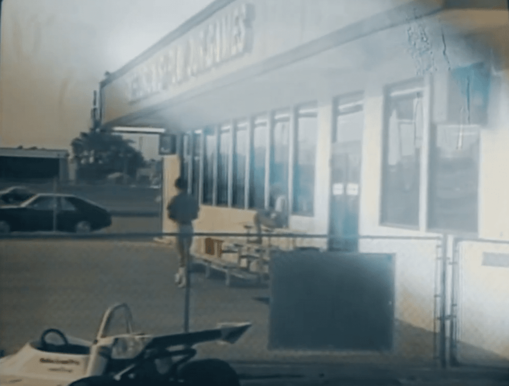 A grainy image of a storefront with the words 'Racers' repeated, showing a person walking near the entrance, a parked car, and a go-kart in the foreground.