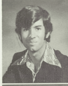 A black and white portrait of a young man with long hair, wearing a patterned shirt and a suit jacket.