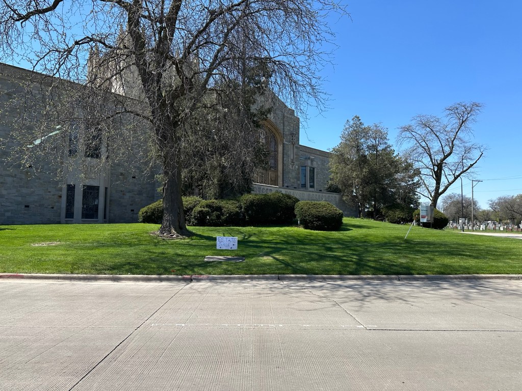 View of a cemetery with a large stone building and well-manicured grassy area, featuring a sign that reads 'Estate Sale'.