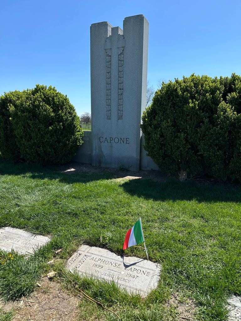Grave site of Al Capone with a large grey headstone marked 'CAPONE' and a smaller headstone for Alphonse Capone, alongside a small Italian flag.