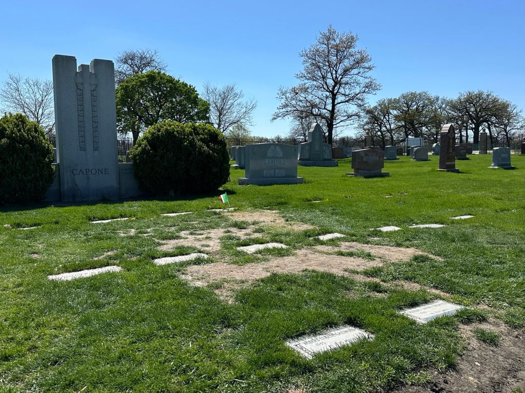 Grave site at Mount Carmel Cemetery featuring a large grey headstone marked 'Capone' surrounded by other gravestones and greenery on a sunny day.