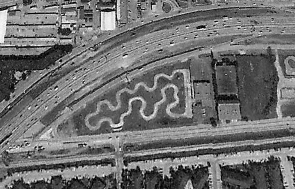 Aerial view of a winding path in a park adjacent to a highway, surrounded by buildings and trees.