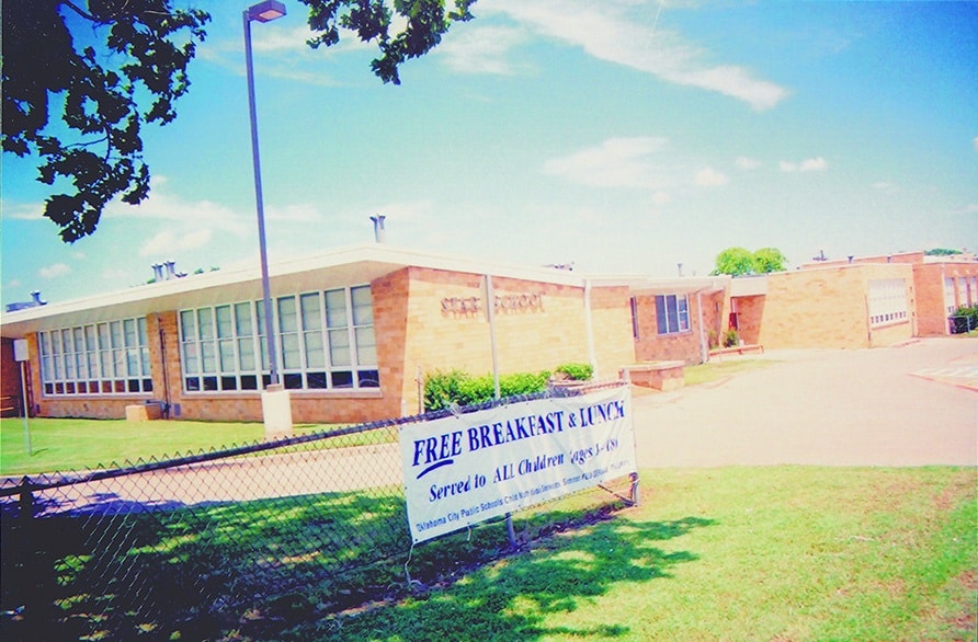 Exterior view of a school building with a sign stating 'FREE BREAKFAST & LUNCH' in front, set against a clear blue sky.