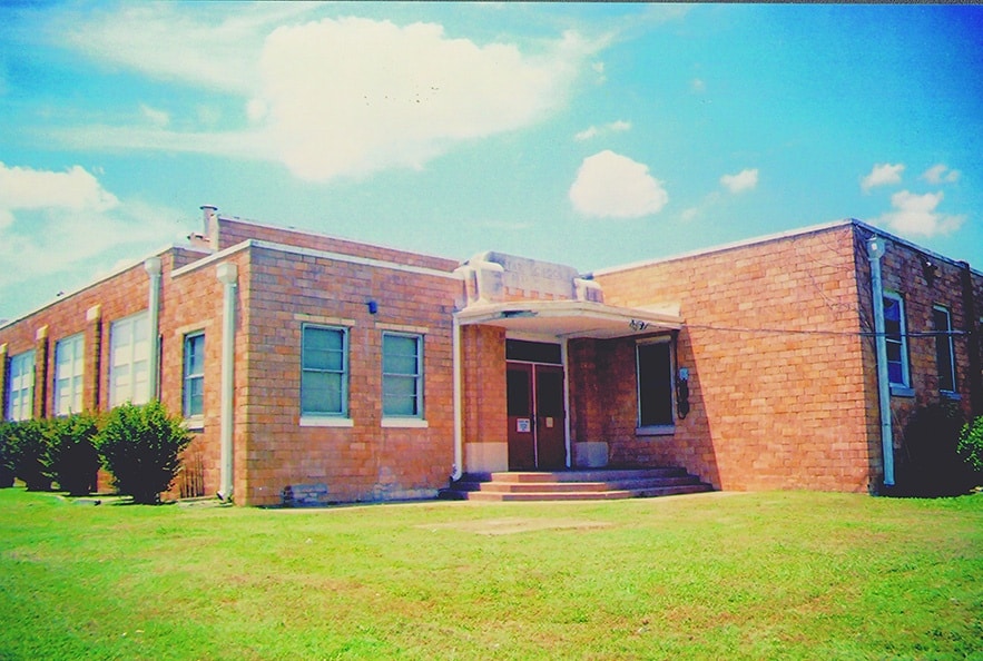 A historical building with a beige stone exterior, featuring large windows and a prominent entrance with steps, surrounded by green grass and a clear blue sky.