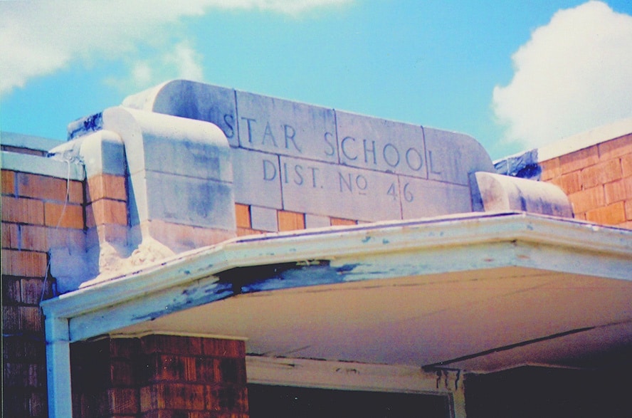 Close-up view of a weathered school building facade with the inscription 'STAR SCHOOL DIST. NO 46' on a stone sign, under a bright blue sky.