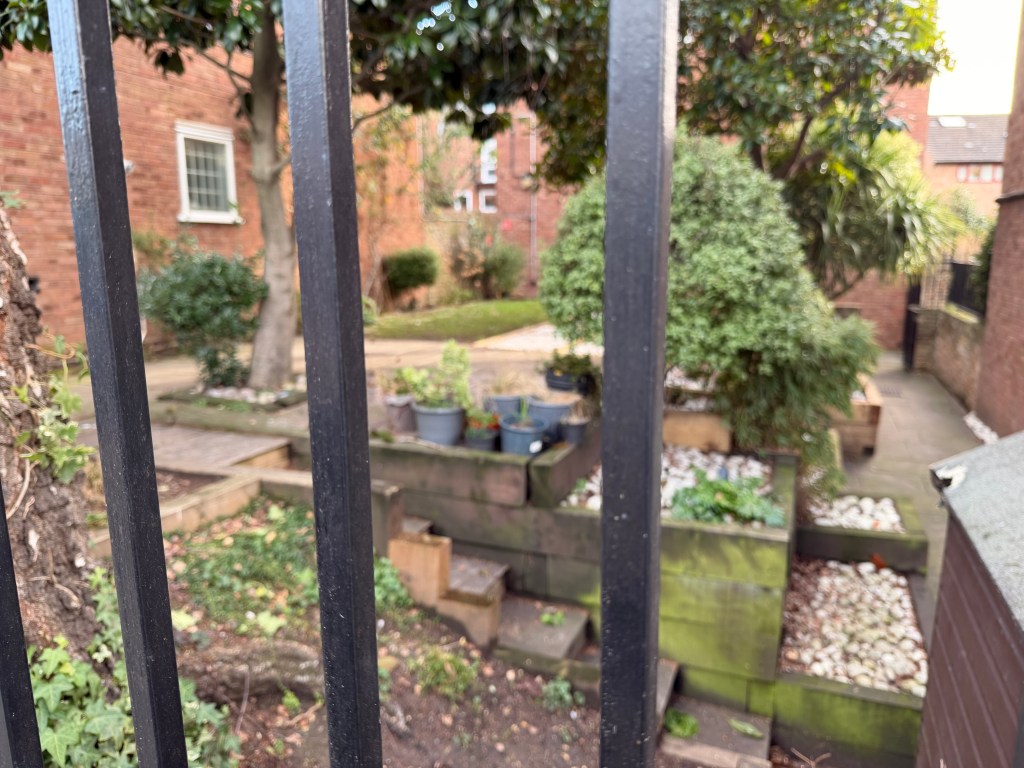 View of a garden through black wrought iron bars, featuring plants in pots, stone paths, and a variety of greenery.