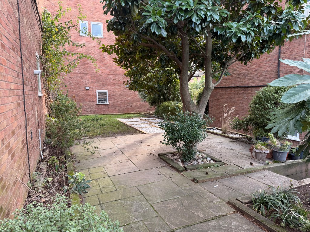 A view of a small garden with a stone patio, surrounded by low shrubs and trees, next to a brick building.