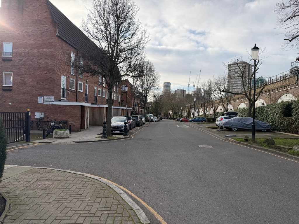 A quiet residential street with brick buildings and parked cars, featuring trees lining the road and modern buildings in the background under a cloudy sky.