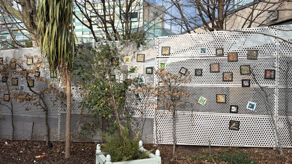 A patterned wall with various framed art pieces displayed, surrounded by greenery and trees in an outdoor setting.