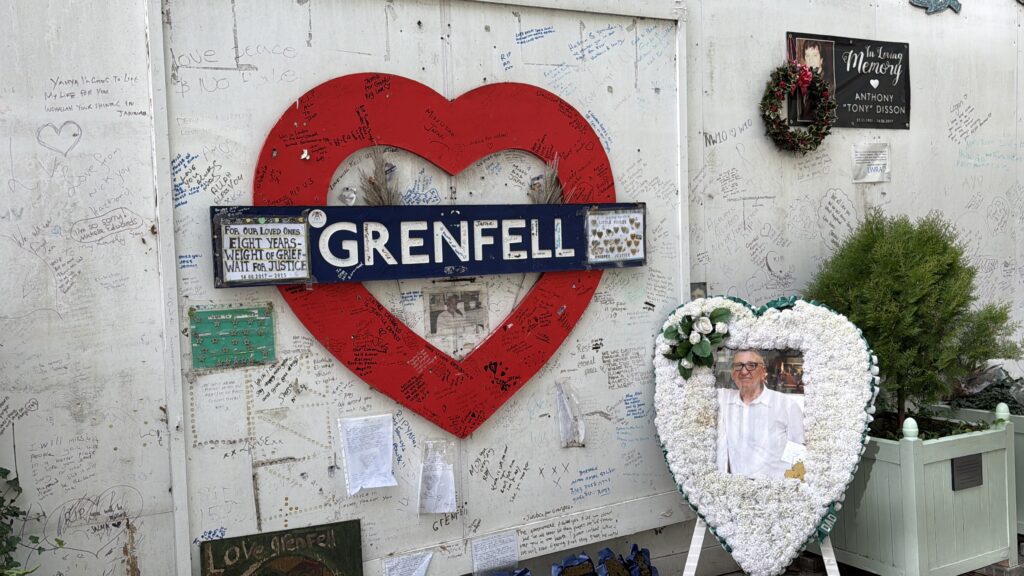 A memorial wall with a large red heart shape and a blue 'Grenfell' sign. Surrounding it are messages and tributes written on the surface. A floral heart-shaped tribute with a framed photo of an older man is displayed in front, along with a wreath and additional messages of remembrance.