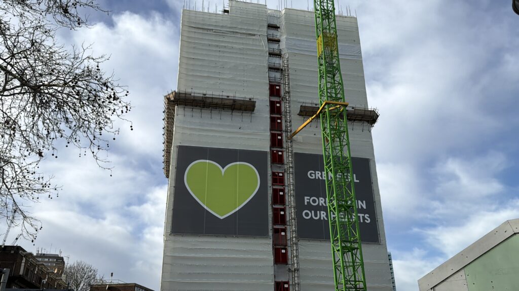 A partially constructed building covered in scaffolding with a large green heart graphic and the text 'Grenfell Forever in Our Hearts' displayed on the side.