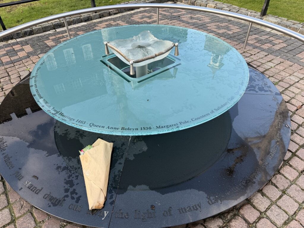 A round memorial with a glass top feature, displaying engraved names and dates of historical figures executed, surrounded by a stone path. A bouquet of flowers rests on the surface.