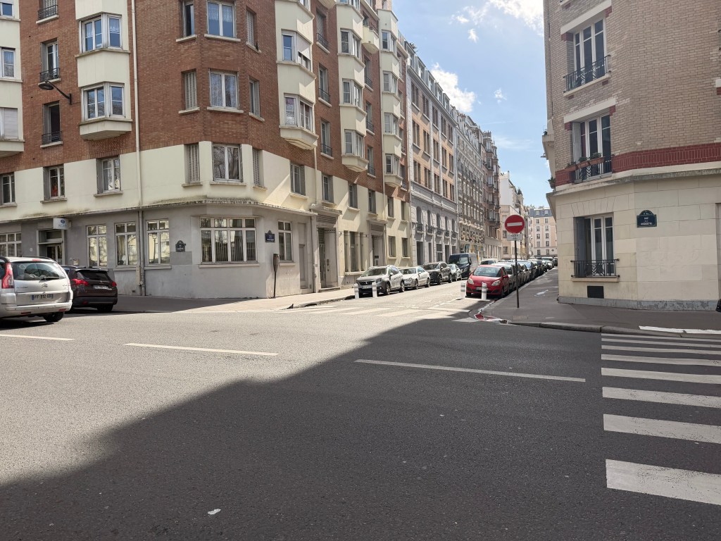 A street view showing a fork in the road with red and brown brick buildings on either side, parked cars along the street, and clear blue skies.