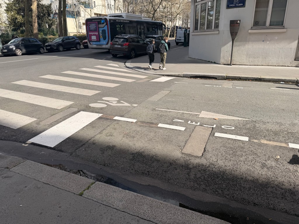 A street scene featuring a crosswalk with pedestrians walking, a city bus in the background, and bicycle lane markings visible on the road.