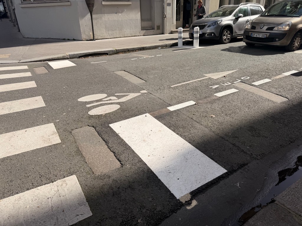 A street corner showing a bike lane symbol on the pavement, a crosswalk with white stripes, and parked cars nearby.