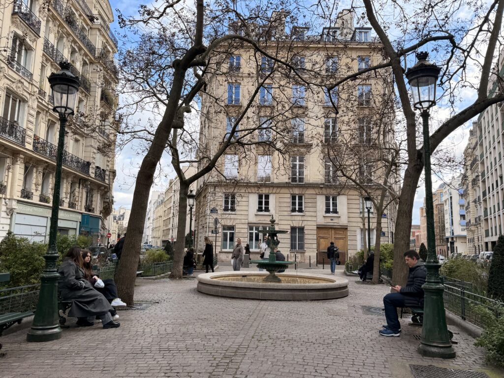 A peaceful Parisian square featuring a central fountain, surrounded by trees and elegant buildings. People are seated on benches and walking by, with a clear sky in the background.